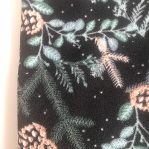 May include: Dark fabric with a winter design. The pattern includes pale green and blue leaves, beige pine cones, and tiny white dots. The material has a soft, textured feel, ideal for seasonal crafts and clothing.