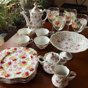 May include: A collection of vintage floral china, including a teapot, cups, saucers, plates, a serving dish, and wine glasses. The set features a white background with red and yellow roses. The plates are square-shaped.