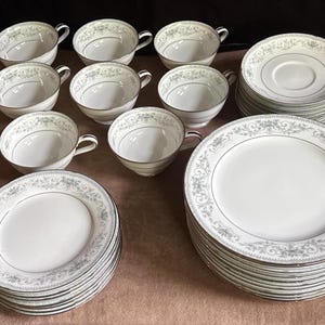 May include: A collection of white porcelain dinnerware with a floral design and silver trim. The set includes teacups, saucers, and plates. The plates and cups are stacked, showcasing the full set. The floral pattern is in shades of gray.