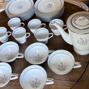 May include: A vintage porcelain tea set with a light blue rim and a floral design. The set includes cups, saucers, plates, and a teapot. The cups and teapot have a silver trim. The set is arranged on a wooden table.