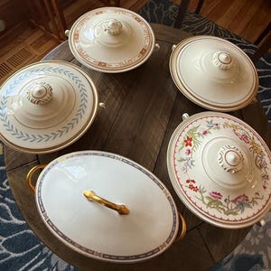 May include: A collection of antique serving dishes with lids, featuring various decorative patterns. The dishes are primarily white with gold trim and handles. Some lids have floral designs, while others have geometric or leaf patterns. The oval dish has a gold handle.