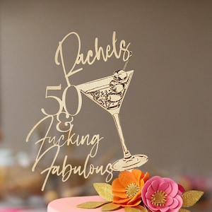 May include: A two-tier cake decorated with pink and teal frosting, gold lace trim, and fabric flowers. A gold cake topper features a martini glass graphic and the text "Rachels 50 & Fucking Fabulous" in a cursive and sans-serif font combination.