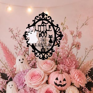 Op de afbeelding: Een zwart decoratief bord met een sierlijke lijst met de tekst A LITTLE BOO IS ALMOST DUE, een witte geest, een spinnenweb, een kat en een pompoen. Het bord staat in een bloemstuk van roze rozen, hortensia's en kleine pompoenen.