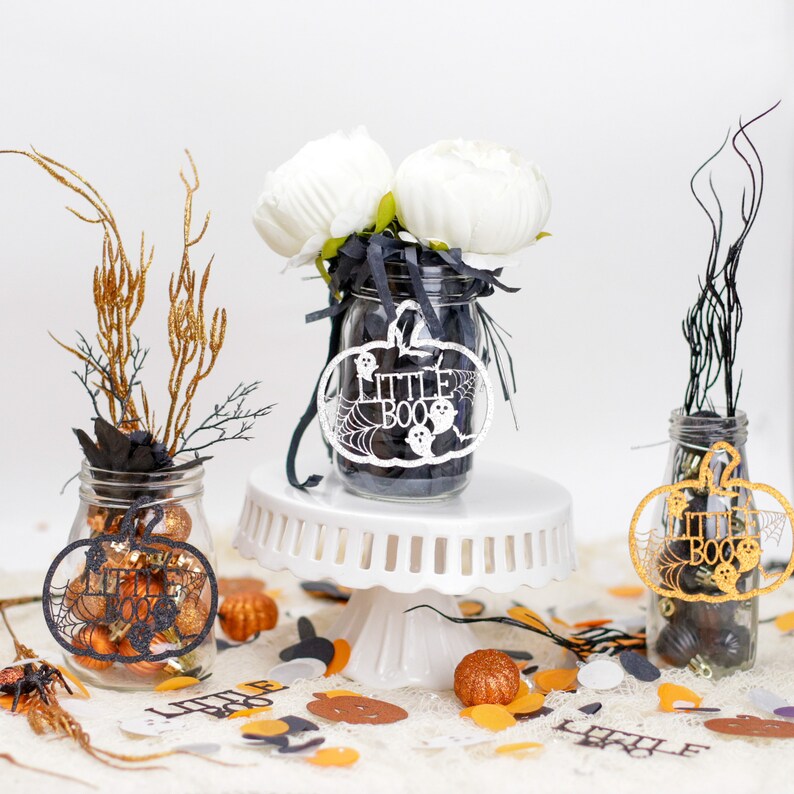 May include: Three glass jars decorated with black and gold Halloween-themed pumpkins and the words "Little Boo". The jars are filled with black and gold glitter and small decorative items. The jars are sitting on a white cake stand and a white surface with orange and black confetti.