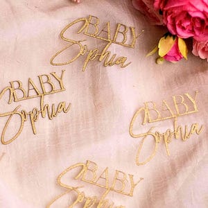 May include: Gold glittered laser-cut word art pieces that spell "BABY Sophia". The script is in a delicate font. The pieces are scattered on a light pink surface, with pink flowers in the background. Ideal for baby shower decorations.