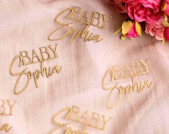 Op de afbeelding: Gouden, glitterende, lasergesneden woordkunststukken met de tekst "BABY Sophia". Het script is in een sierlijke lettertype. De stukken zijn verspreid op een lichtroze oppervlak, met roze bloemen op de achtergrond.