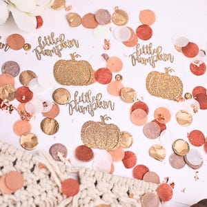 May include: Gold glitter confetti with the words "little pumpkin" and a pumpkin design. The confetti is scattered on a white background with other confetti in shades of peach, brown, and gold.