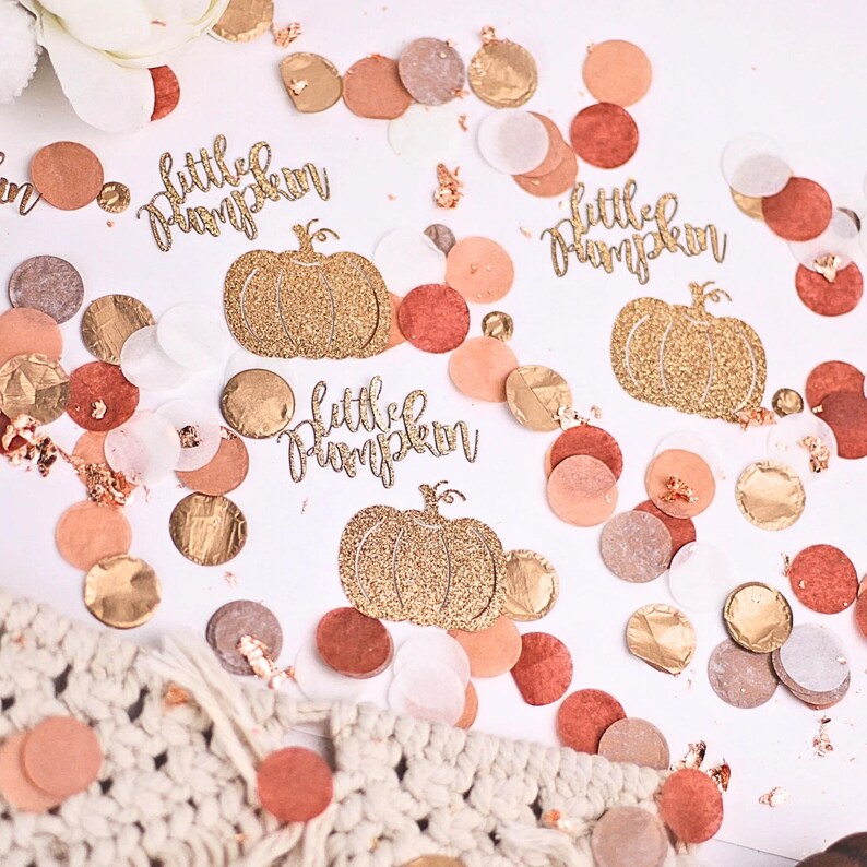 May include: Gold glitter confetti with the words "little pumpkin" and a pumpkin design. The confetti is scattered on a white background with other confetti in shades of peach, brown, and gold.