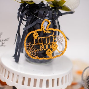 May include: A gold glitter pumpkin shaped ornament with the words "Little Boo" and a spider web design.