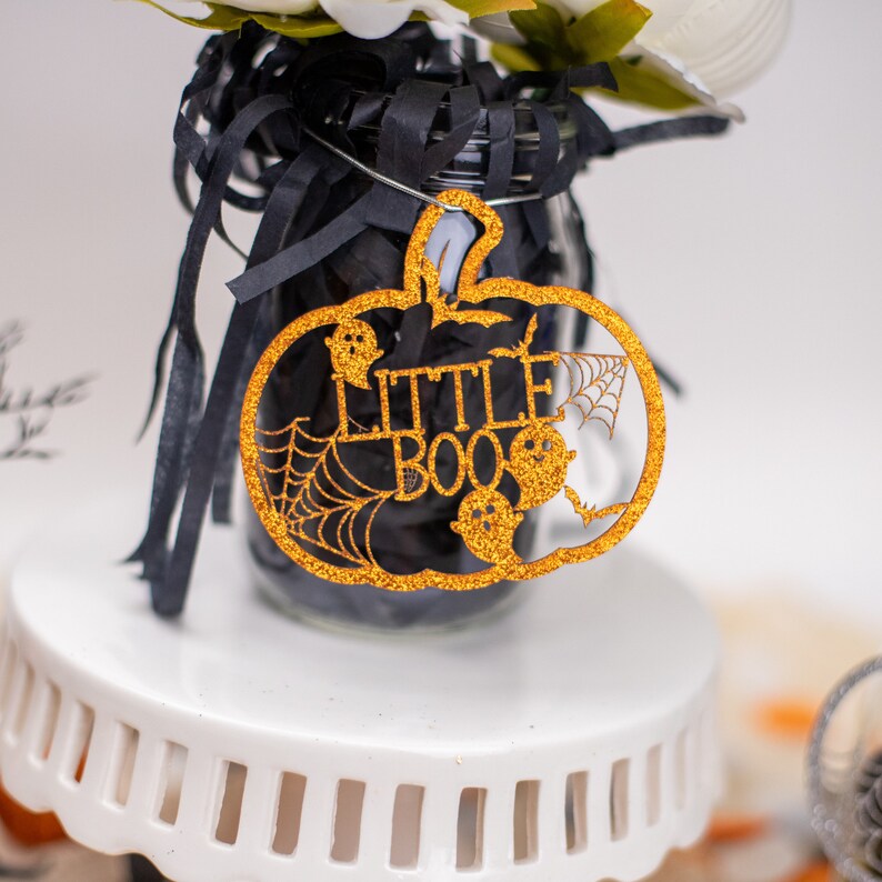 May include: A gold glitter pumpkin shaped ornament with the words "Little Boo" and a spider web design.
