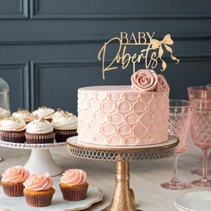 Pode incluir: Um bolo rosa e branco com um topo de bolo dourado que diz "Baby Roberts" com um laço dourado. O bolo está em um suporte de bolo dourado e há cupcakes em um suporte de bolo branco na frente dele.