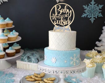 May include: A two-tiered cake with light blue and white frosting, decorated with snowflakes. A wooden cake topper reads "Baby it's cold outside." Cupcakes and cookies are also on display, creating a winter-themed dessert table.