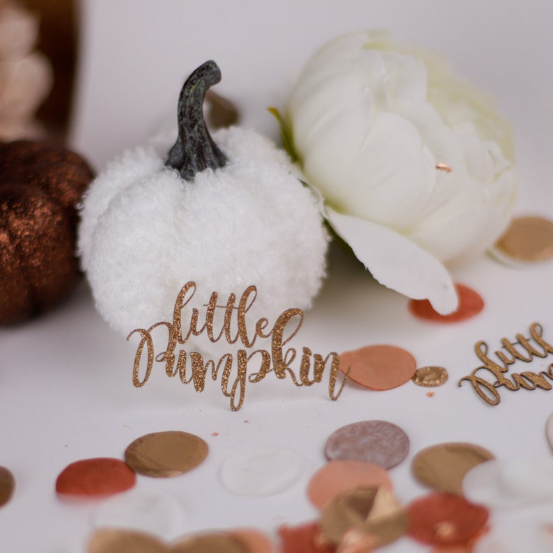 May include: A white faux fur pumpkin with a brown stem sits on a white surface with gold glitter confetti and a gold glitter sign that says "little pumpkin".
