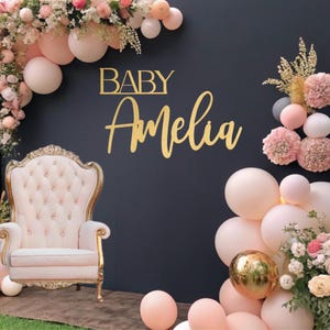May include: A pink and gold baby throne chair with a black backdrop decorated with pink and white balloons, flowers, and the text "BABY Amelia".