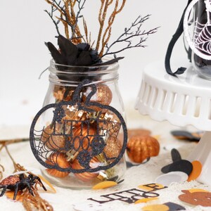 May include: A glass jar filled with orange and gold glitter ornaments and a black pumpkin cutout with the text "Little Boo" hanging from the top. The jar is decorated with black feathers and gold branches.