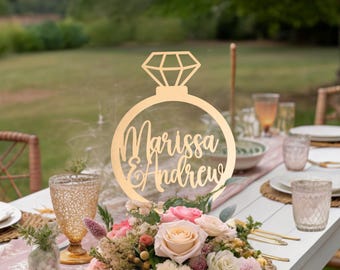 Puede incluir: Un topper de mesa de madera con forma de anillo y diamante con los nombres "Marissa & Andrew" escritos en cursiva. El topper está colocado sobre una mesa con un centro de mesa floral.