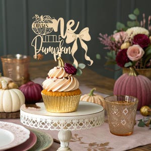 May include: A cupcake with peach-colored frosting and a wooden cake topper that reads "Olivia's Little Pumpkin." The cupcake sits on a white, ornate cake stand. The background includes pumpkins and flowers.