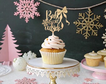 May include: A cupcake with pink frosting and a wooden cake topper that reads "Baby it's cold outside". The topper has a bow and snowflake design. The cupcake sits on a white and gold cake stand, with other cupcakes in the background.