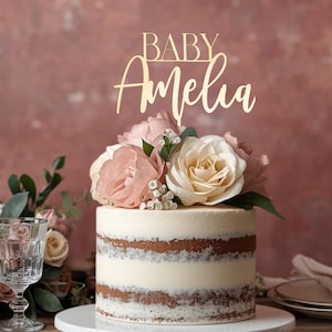 May include: A layered cake with white frosting and visible cake layers, topped with pink and cream-colored roses and a gold-colored "BABY Amelia" cake topper. The cake sits on a white cake stand, with a glass and flowers in the background.