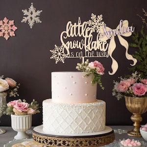 Könnte beinhalten: Eine zweistöckige Torte mit hellrosa Oberteil und weißem Boden, verziert mit einem goldenen Tortenaufsatz mit der Aufschrift "A little snowflake is on the way" und dem Namen "Amelia". Der Kuchen steht auf einem goldenen Ständer, umgeben von Blumenarrangements und dekorativen Schneeflocken.