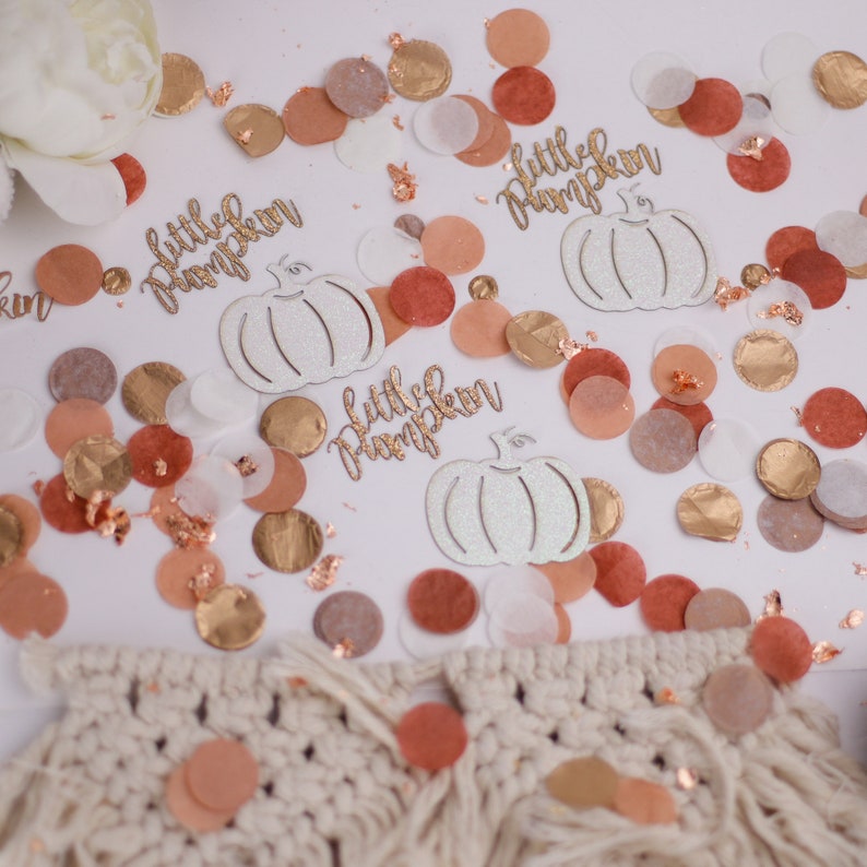 May include: White and gold confetti with the text "little pumpkin" on three of the pieces. The confetti is scattered on a white surface with a white and beige crocheted piece in the bottom of the image.