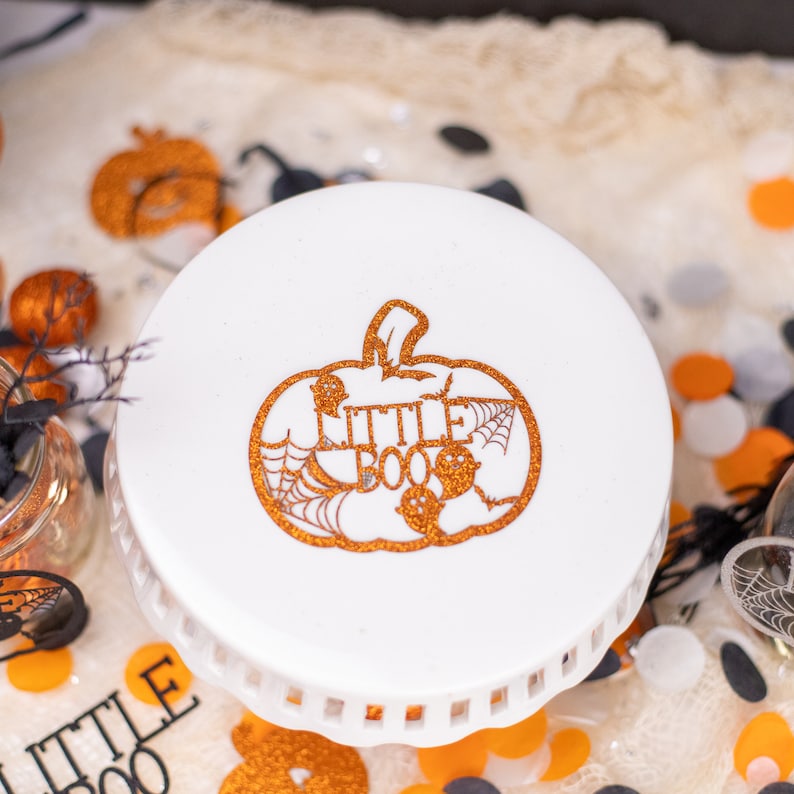 May include: White cake stand with a scalloped edge. The top of the stand has a pumpkin design with the words "Little Boo" in orange glitter.