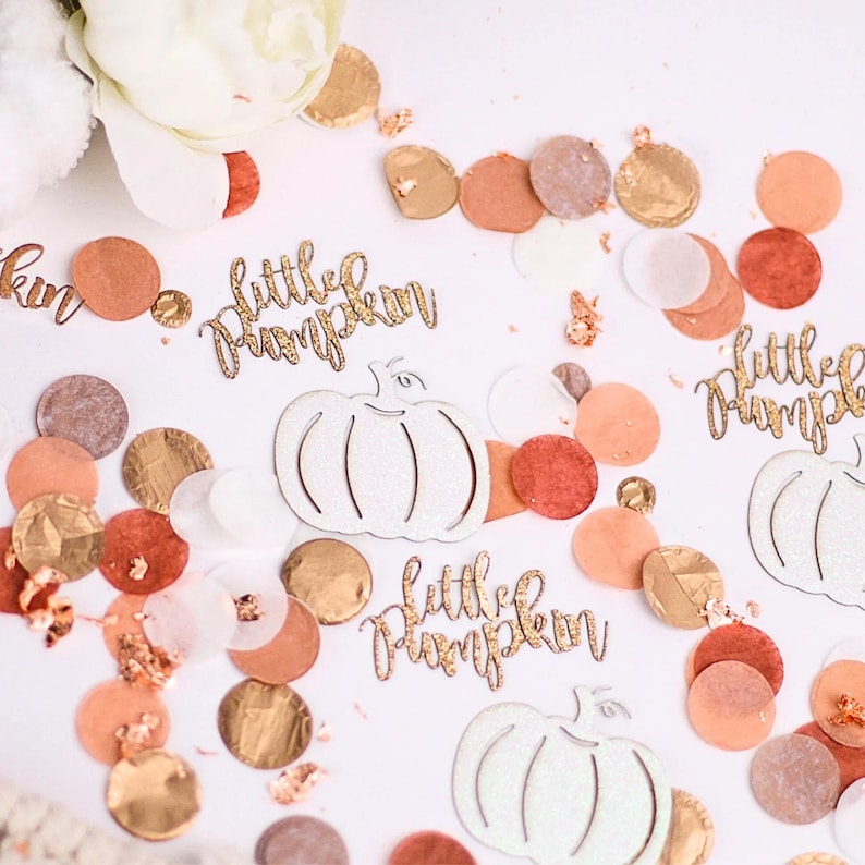 May include: White glitter pumpkin cutouts with the text "little pumpkin" in gold glitter. The cutouts are scattered on a white surface with peach, brown, and gold confetti.
