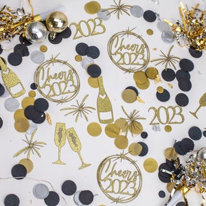 Peut inclure: Confettis dorés et noirs avec des décorations dorées scintillantes 2023, y compris une bouteille de champagne, des flûtes à champagne et une bannière 'Cheers 2023'.