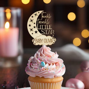 May include: A cupcake topped with pink frosting and sprinkles, with a wooden topper that reads "Twinkle Twinkle Little Star Baby Olivia" in script. The cupcake sits on a white plate, with a lit candle and blurred background.
