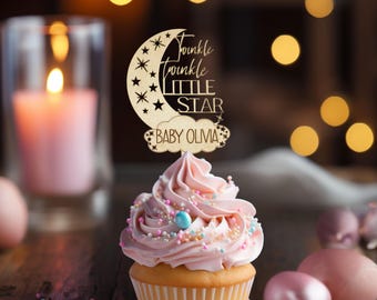 May include: A cupcake topped with pink frosting and sprinkles, with a wooden topper that reads "Twinkle Twinkle Little Star Baby Olivia" in script. The cupcake sits on a white plate, with a lit candle and blurred background.