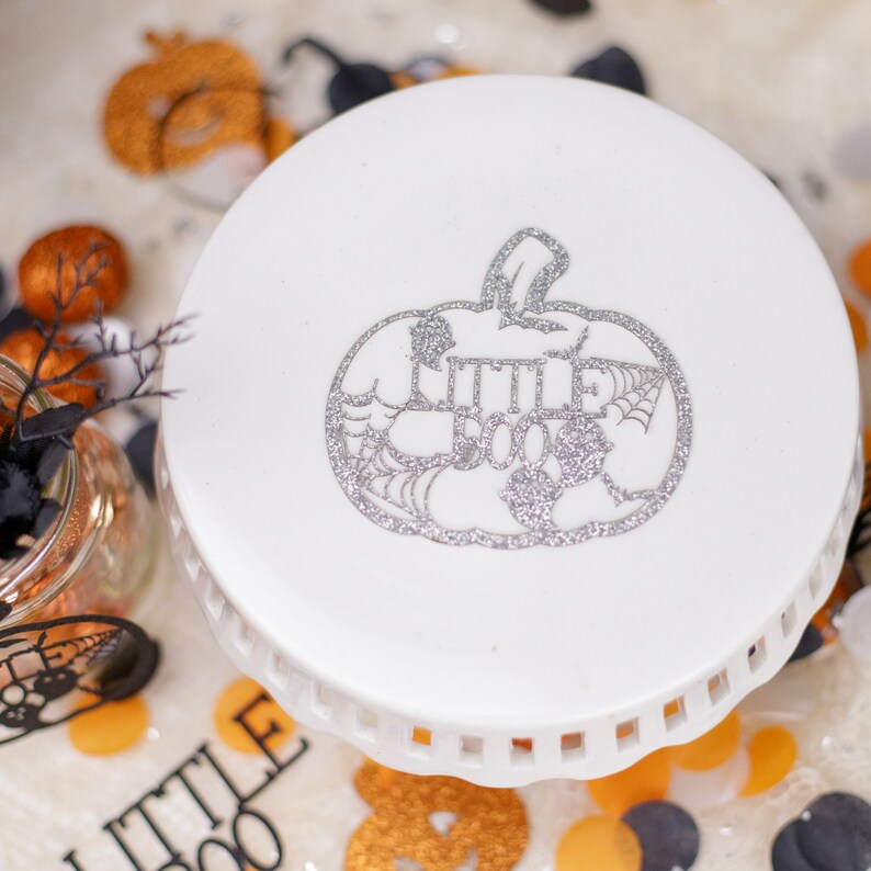 May include: White ceramic cake stand with a silver glitter pumpkin design that says "Little Boo" on it. The stand is decorated with orange and black Halloween sprinkles.