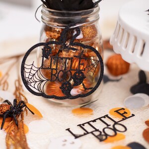 May include: Black pumpkin-shaped Halloween decoration with the words "Little Boo" inside a spiderweb design. The decoration is hanging from a glass jar filled with orange and gold glitter ornaments.