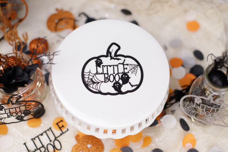 May include: White cake stand with a black and white pumpkin design. The pumpkin has a spiderweb and the words "Little Boo" inside.