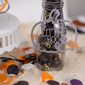 May include: A clear glass jar filled with small black and silver objects, decorated with a silver glitter pumpkin cutout that says "Little Boo".