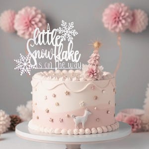 Könnte beinhalten: Ein weißer Kuchen mit rosa Zuckerguss und einem weißen Kuchentopper mit der Aufschrift "A little snowflake is on the way".