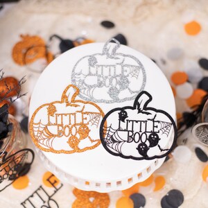 May include: Three pumpkin-shaped Halloween decorations with the text "Little Boo" in black and white glitter. The pumpkins are on a white cake stand.