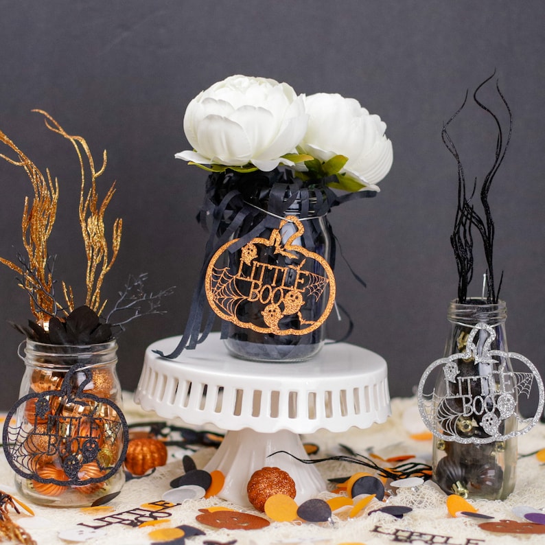 May include: Three glass jars decorated for Halloween with black and gold glitter, black and white spiderwebs, and the words "Little Boo". The jars are filled with black and orange candy and are sitting on a white cake stand. The jars are surrounded by black and orange confetti.