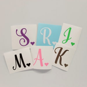 May include: A collection of personalized decals featuring various letters of the alphabet. Each letter, in colors like purple, pink, green, and brown, is accompanied by a small heart. The letters are displayed on white square backgrounds.