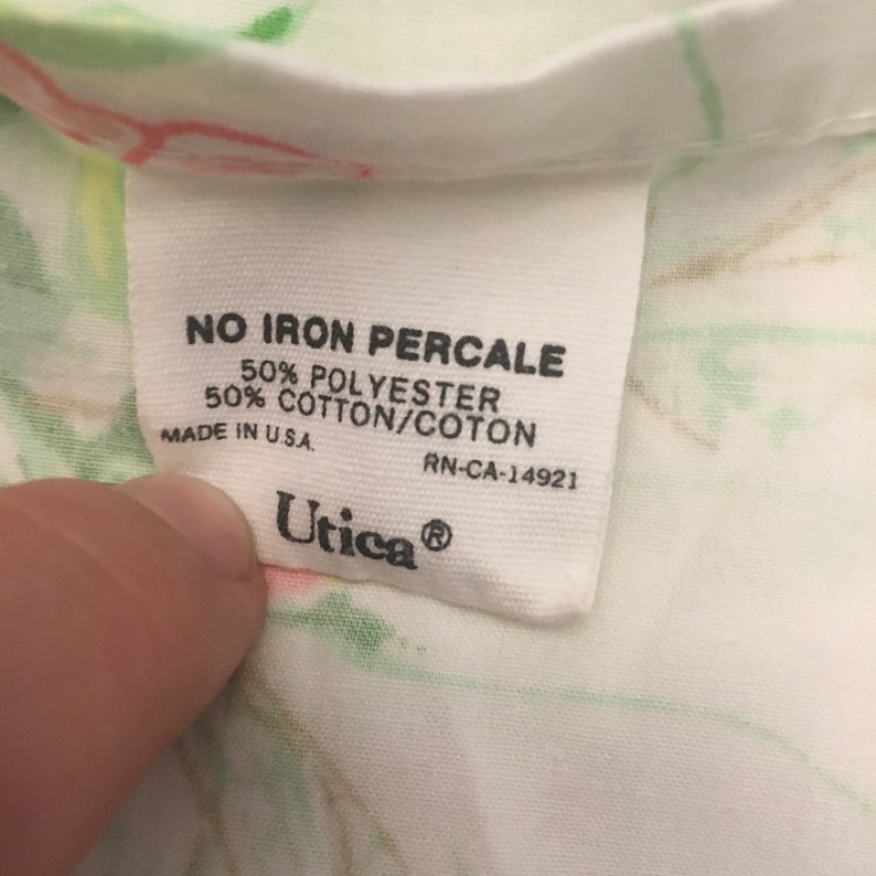 Puede incluir: Primer plano de una etiqueta de tela blanca con texto negro. La etiqueta dice "NO IRON PERCALE", "50% POLYESTER", "50% COTTON/COTON", "MADE IN U.S.A." y "Utica&reg;". La etiqueta est&aacute; sobre una tela blanca con estampado floral.