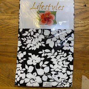 May include: A packaged tablecloth with a black and white floral pattern. The tablecloth is labeled "Lifestyles" with a floral graphic. The dimensions are 60 x 102 inches, suitable for seating 8-10 people.