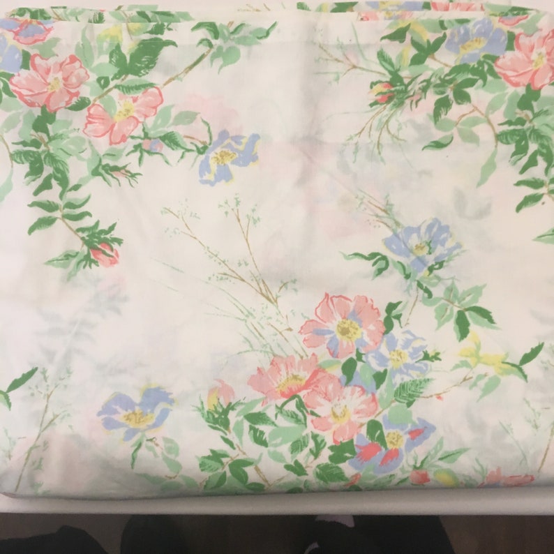 Puede incluir: S&aacute;bana blanca con un dise&ntilde;o floral que presenta flores rosas, azules y amarillas con hojas verdes. El patr&oacute;n floral se repite por toda la superficie de la tela.