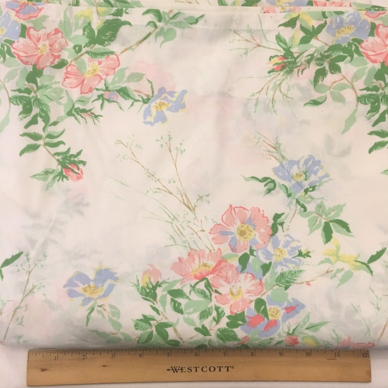 Puede incluir: Tela blanca con un estampado floral de flores rosas, azules y amarillas con hojas y tallos verdes. Las flores est&aacute;n esparcidas por la tela. Una regla de madera en la parte inferior.