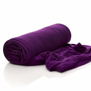 May include: A rolled-up, deep purple blanket next to a portion of the unfolded blanket. The fabric appears soft and textured. The blanket is on a white surface.