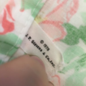 Puede incluir: Primer plano de una tela estampada con flores rosas y verdes sobre un fondo blanco. Una etiqueta dice "J.P. Stevens & Co. Inc. &copy; 1978". La tela es probablemente vintage y adecuada para acolchar o manualidades.