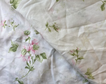 May include: White fabric with a floral pattern featuring pink flowers and green leaves. The fabric is folded, showing the pattern on both sides. The flowers have a watercolor effect, and the leaves are a muted green.