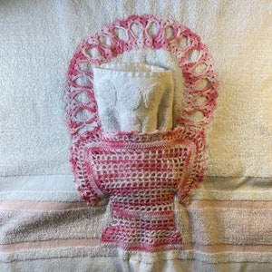 May include: A white towel with a decorative pink and white crocheted pocket and trim. The pocket holds a folded white towel. The crocheted trim has a scalloped edge and a geometric pattern. The towel has pink stripes.