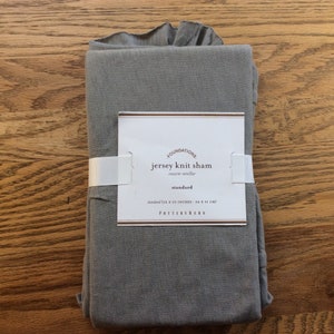 May include: A folded gray jersey knit sham, standard size 26 x 20 inches, with a white label and ribbon. The label reads "FOUNDATIONS jersey knit sham" and "POTTERY BARN". The sham is on a wooden surface.