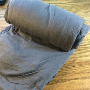 May include: A rolled-up, gray fabric item, possibly a blanket or clothing, rests on a wooden surface. The fabric appears soft and has a slightly wrinkled texture. The item is folded over itself.