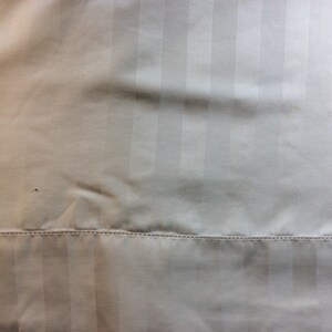 May include: Close-up of a cream-colored fabric with vertical stripes. The fabric has a subtle sheen and a stitched seam at the bottom. The texture appears smooth and slightly wrinkled.