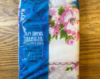 May include: Sealed package of two standard size pillowcases. The pillowcases are white with a floral pattern in pink and green. The packaging is blue and features text that reads "No Iron Muslin Pillowcases". The pillowcases are 20x26 inches.